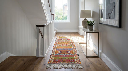 A colorful patterned runner rug with fringe is displayed on a wooden floor in a bright, modern hallway with a staircase, console table, and lamp.