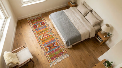A colorful geometric patterned runner rug with fringe is styled on a light wood floor in a modern bedroom setting next to a bed with neutral bedding.