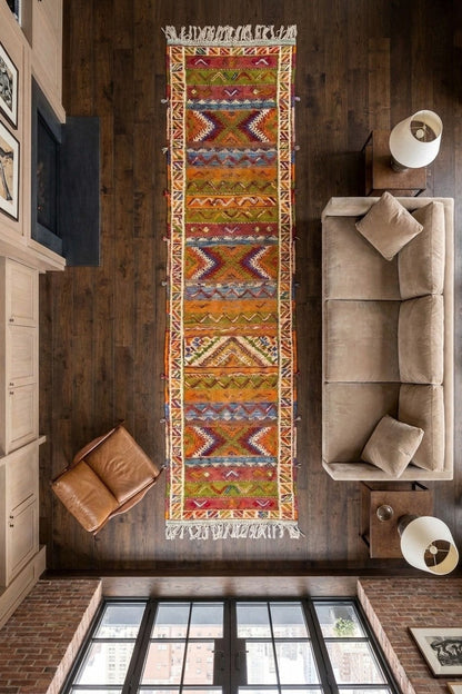 A long rectangular area rug with a colorful geometric tribal pattern is shown from a top-down view in a modern living room setting with dark wood floors, a beige sofa, and a brown leather chair.
