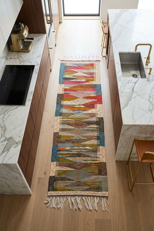 A long, narrow geometric patterned runner rug with fringe ends laid out on a light wood floor in a modern kitchen setting with marble countertops and wooden cabinets.