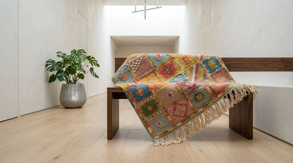 A colorful patchwork-style moroccan rug  draped over a wooden bench in a bright, modern room with light wood floors and a potted plant.