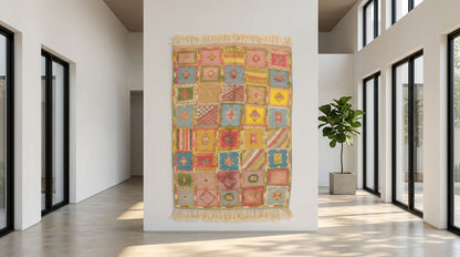A colorful patchwork-style woven moroccan rug with gold fringe hangs on a white wall in a bright, modern hallway with large black-framed windows and a potted plant.
