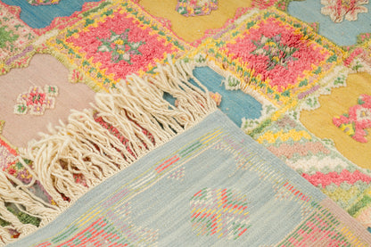 A close-up of a colorful geometric moroccan rug with cream fringe, featuring geometric and floral patterns in pink, yellow, blue, and beige.
