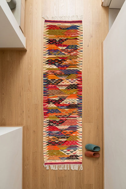 A long, narrow woven runner rug with a colorful geometric pattern hangs on a light wood floor in a minimalist interior. The rug features a cream base with repeating diamond and zigzag motifs in red, orange, blue, green, and yellow.