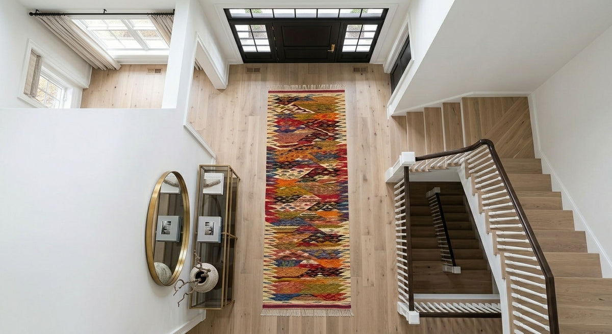 A long, multicolored patterned runner rug is displayed on a light wood floor in a bright, modern home entryway with a staircase, mirror, and console table.