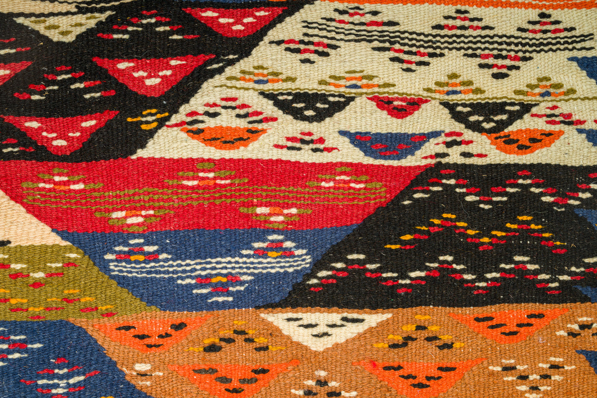 A close-up of a woven kilim textile with a colorful geometric pattern featuring triangles and dotted motifs in red, blue, orange, black, beige, and olive green.