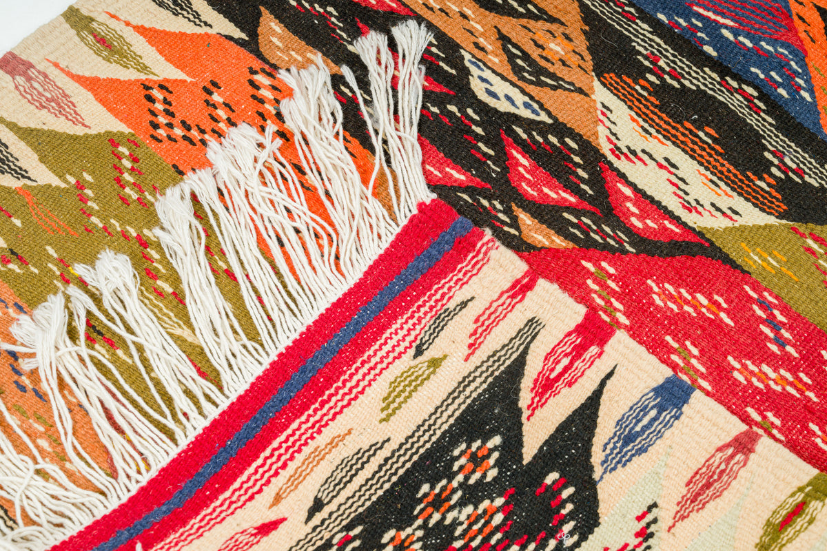 A close-up of a woven textile with geometric patterns in red, orange, black, beige, blue, and olive green, featuring white fringe on one side.