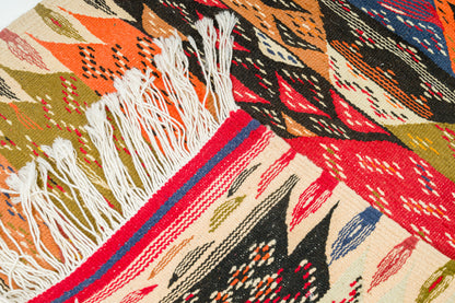 A close-up of a woven textile with geometric patterns in red, orange, black, beige, blue, and olive green, featuring white fringe on one side.