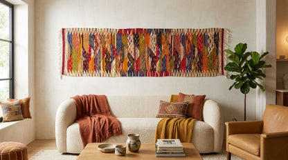 A large, vibrant bohemian-style woven wall tapestry with intricate geometric patterns in red, orange, yellow, blue, green, and brown, hanging on a cream-colored wall above a modern white sofa in a styled living room.