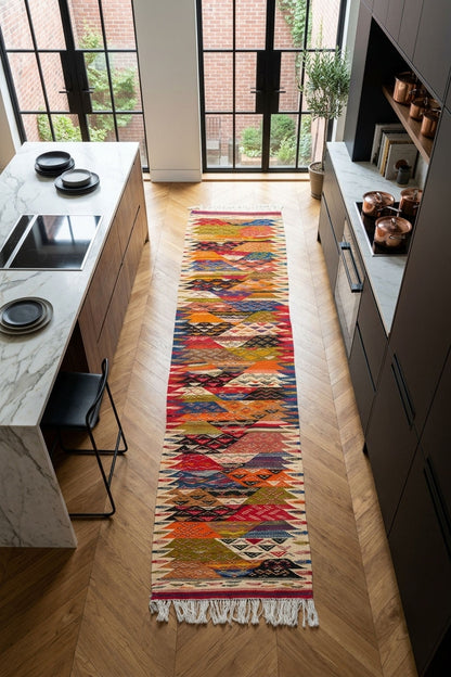 A long, colorful geometric patterned runner rug with white fringe is displayed on a light wood herringbone floor in a modern kitchen setting.
