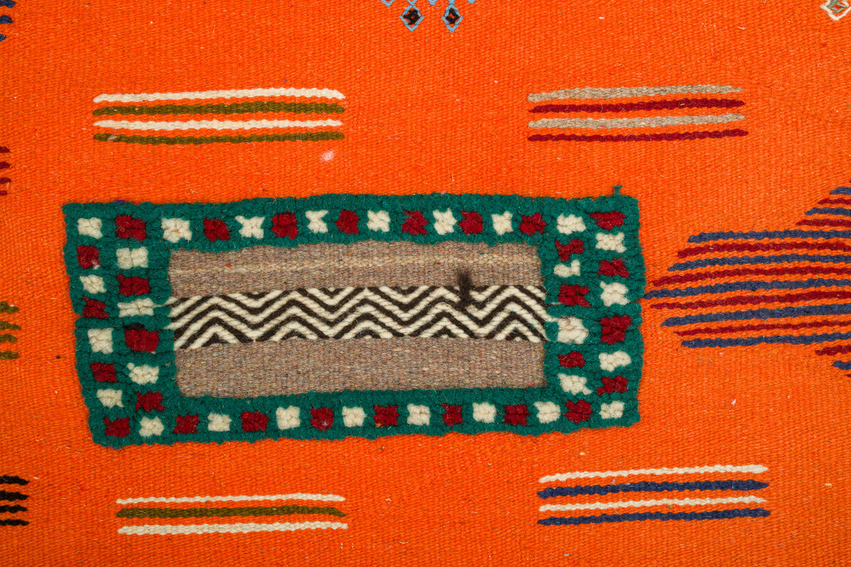 Close-up of a Moroccan Rug with a bright orange base, featuring a central rectangular motif in teal, beige, gray, and red, surrounded by small striped accents. 