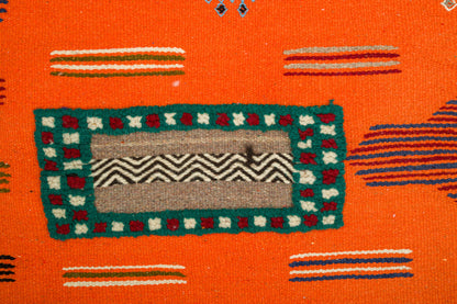 Close-up of a Moroccan Rug with a bright orange base, featuring a central rectangular motif in teal, beige, gray, and red, surrounded by small striped accents. 