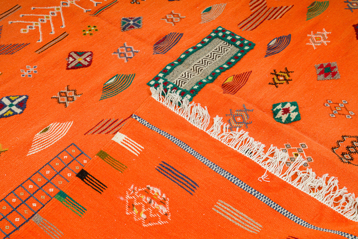 A close-up of a bright orange Moroccan woven rug with geometric patterns, including a green and white patterned rectangular motif, white fringe, and small multicolored decorative symbols.