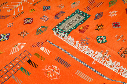 A close-up of a bright orange Moroccan woven rug with geometric patterns, including a green and white patterned rectangular motif, white fringe, and small multicolored decorative symbols.
