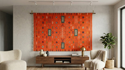 A large orange Moroccan woven tapestry with geometric patterns hangs on a wooden rod in a modern living room setting with a wooden console table, two cream armchairs, and a potted plant.