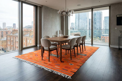 A modern dining room with a large orange patterned rug under a wooden dining table and six upholstered chairs, set in a high-rise apartment with floor-to-ceiling windows and a city skyline view.