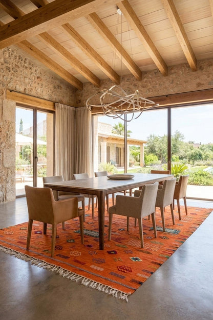 A dining room interior with a rectangular wooden dining table surrounded by beige upholstered chairs, set on a large orange patterned rug. The room features a high wood-beamed ceiling, stone walls, and large windows looking out to a garden.