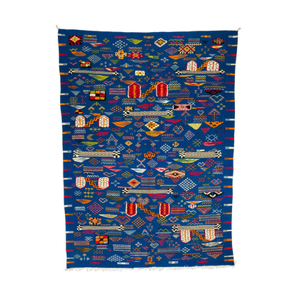 A square blue Moroccan woven rug with a dense, colorful geometric patterns, flags, and abstract shapes, finished with white fringe on the bottom edge.