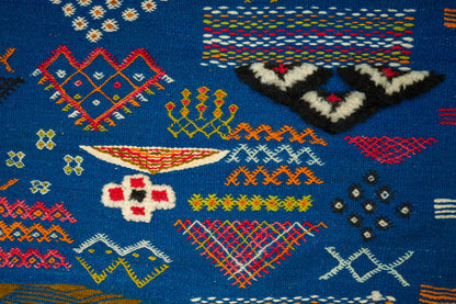 Close-up of a blue moroccan kilim rug with colorful geometric embroidery and motifs including diamonds, zigzags, and abstract shapes in red, yellow, white, black, and orange. 