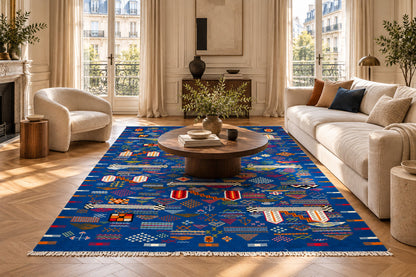 A large rectangular blue kilim area rug with a vibrant blue base and intricate multicolor geometric patterns, styled in a bright, modern living room with a wooden coffee table and cream-colored sofa.