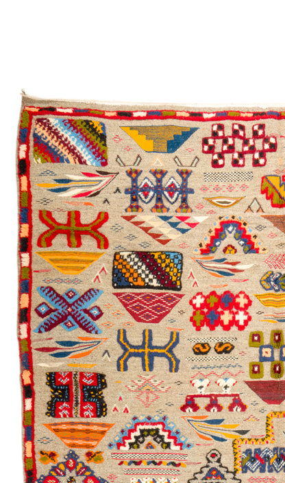 A close-up of a colorful geometric patterned rug with a cream base and red border, featuring various tribal motifs in red, blue, yellow, and green.