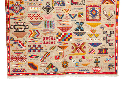 A square handwoven kilim rug with a cream-colored base and a dense collection of colorful geometric and tribal motifs, finished with white fringe on the bottom edge.