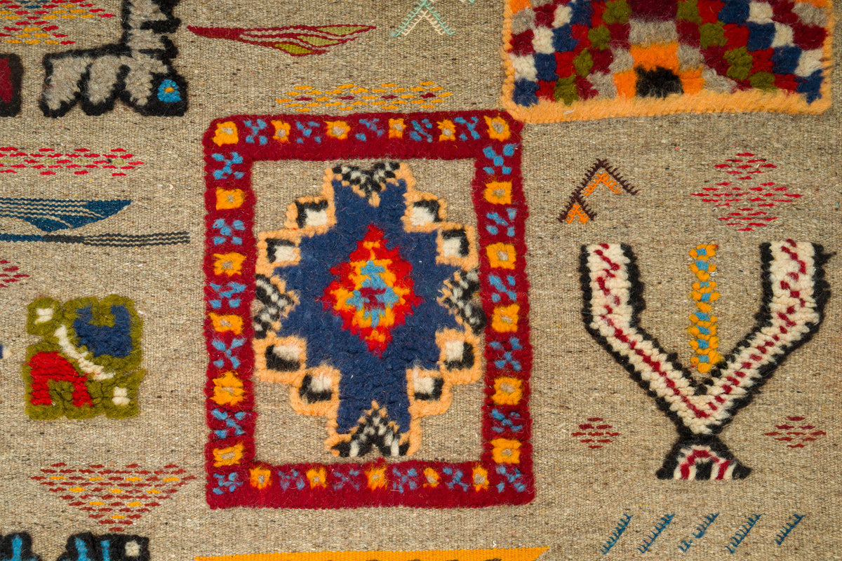 A close-up of a woven rug with a central geometric medallion design in blue, red, orange, yellow, and black on a beige background, surrounded by other colorful tribal-style patterns.