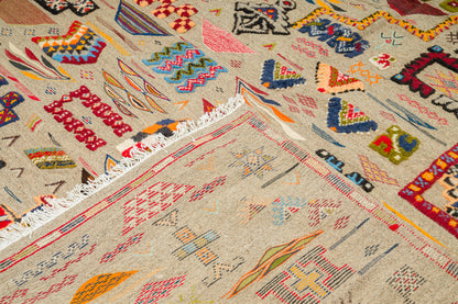 A close-up of a woven rug with a beige base and colorful geometric patterns, including red, blue, orange, green, and black motifs, with white fringe visible along one edge.