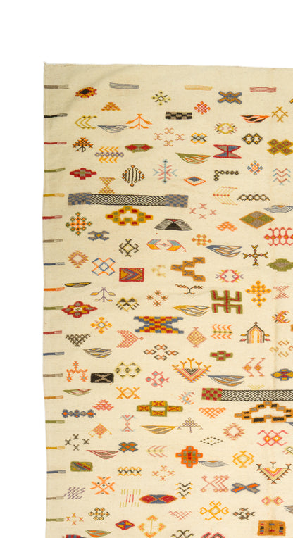 A close-up of a Moroccan rug with a cream base and dense rows of small geometric and symbolic motifs in orange, brown, red, gray, and black.