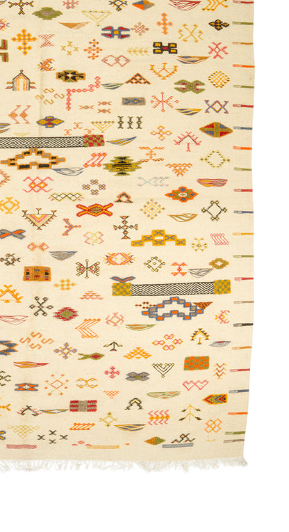 A close-up of a wovenwool rug with a cream background and dense multicolor geometric and tribal-style motifs, including orange, red, yellow, green, and brown elements, with fringe visible along the bottom edge.