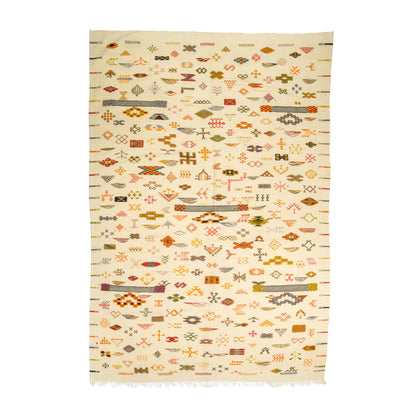 A Moroccan flat weave kilim rug with a cream background and dense geometric patterns in orange, brown, olive green, and pink, finished with short fringe on the bottom edge.