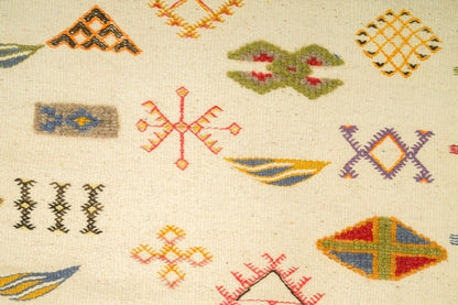Close-up of a woven rug with a cream background and scattered colorful geometric motifs in orange, green, blue, yellow, and purple.