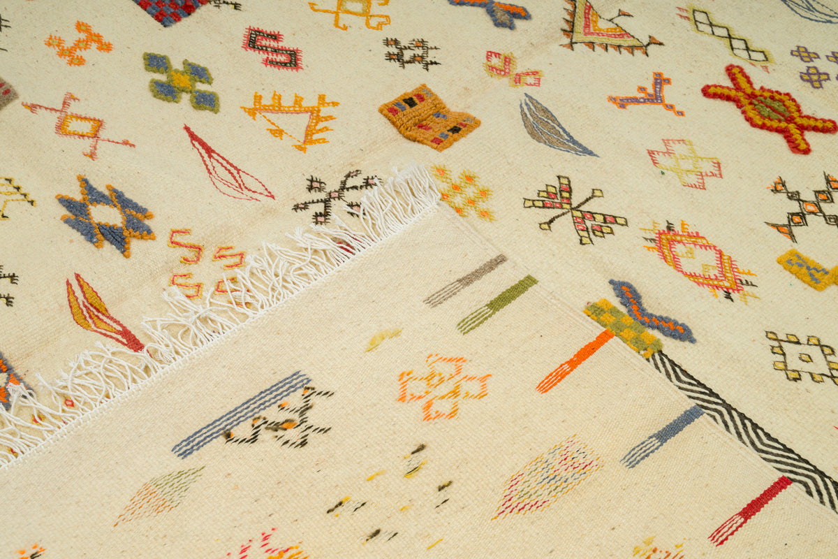 A close-up of a 100% wool rug with a cream base and multicolored geometric motifs, including orange, blue, yellow, green, and red shapes, with white fringe visible along one edge.