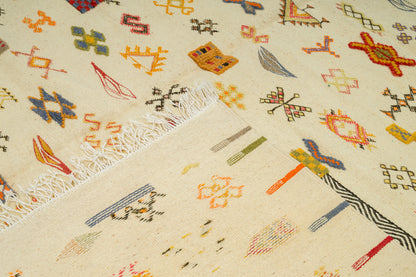 A close-up of a 100% wool rug with a cream base and multicolored geometric motifs, including orange, blue, yellow, green, and red shapes, with white fringe visible along one edge.
