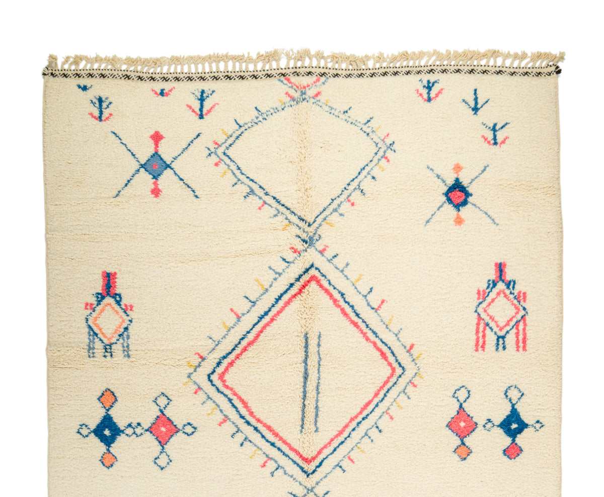 A square cream-colored Moroccan wool rug with a colorful geometric pattern in blue, red, and orange, featuring a central diamond motif and fringed top edge.