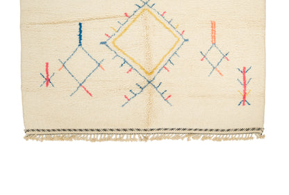 A close-up of a cream-colored Moroccan rug featuring a central yellow diamond motif and blue and red geometric accents, finished with a fringed edge.