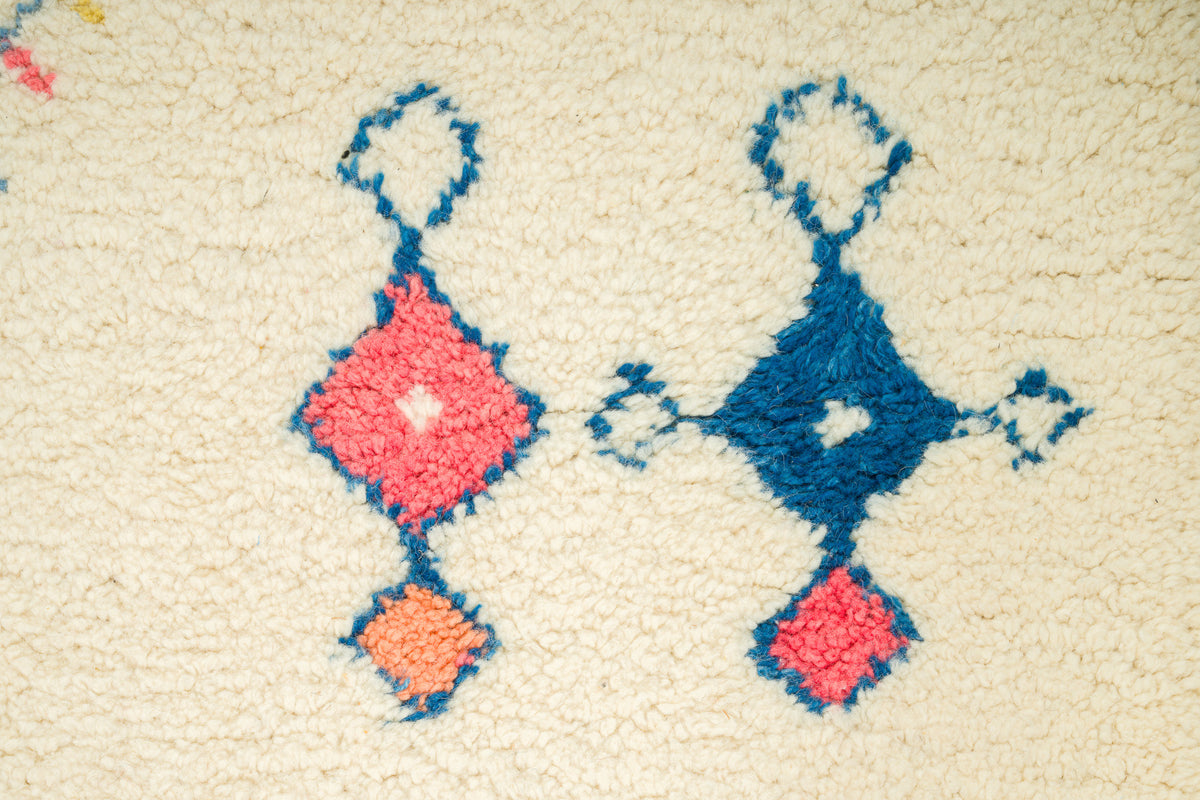 A close-up of a beautiful Moroccan rug, loop-pile rug featuring a traditional Moroccan geometric pattern in blue, coral pink, and orange on a cream-colored background.