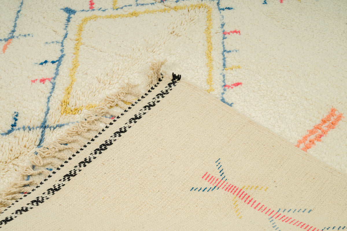 Close-up of a cream-colored woven rug with multicolor geometric lines in blue, yellow, and orange, featuring black and white patterned fringe.