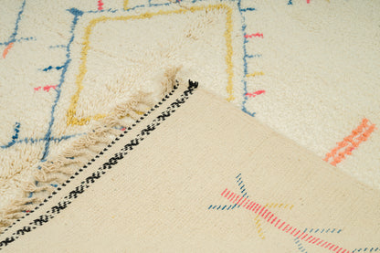 Close-up of a cream-colored woven rug with multicolor geometric lines in blue, yellow, and orange, featuring black and white patterned fringe.