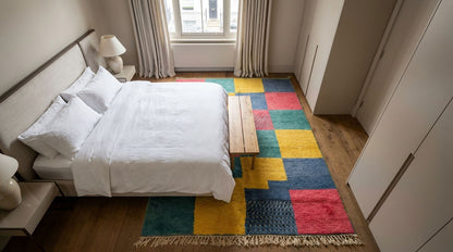 A colorful patchwork area rug with geometric blocks of yellow, blue, red, and green, styled in a modern bedroom next to a bed with white linens.
