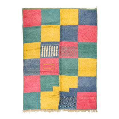 A square multicolor patchwork rug with a woven texture and fringed edges, featuring blocks of teal green, mustard yellow, dusty blue, and coral pink with various geometric patterns.