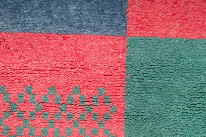 A close-up of a moroccan rug with a checkered pattern in red, blue, and green, featuring a small geometric motif in green on the red section.