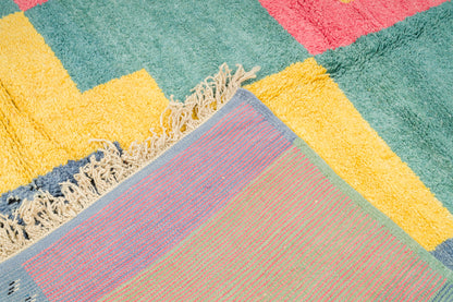 A close-up of a colorful geometric area rug with a chunky shag texture and fringed edge. The design features large blocks of teal green, mustard yellow, dusty pink, and lavender with a woven border.