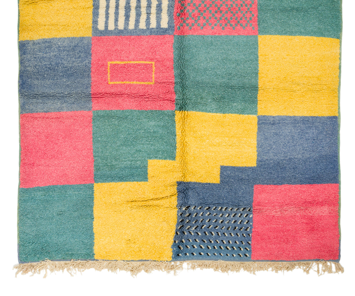 A square rug with a colorful patchwork design, featuring blocks of teal green, mustard yellow, dusty blue, and coral pink with textured geometric patterns and a fringed edge.