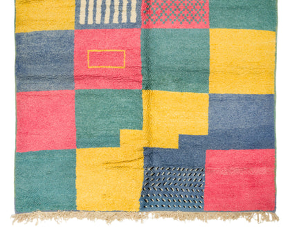 A square rug with a colorful patchwork design, featuring blocks of teal green, mustard yellow, dusty blue, and coral pink with textured geometric patterns and a fringed edge.