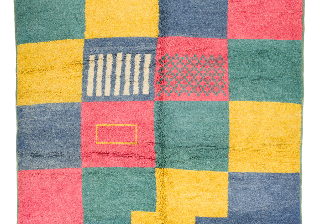 A close-up of a colorful woven textile with a patchwork grid pattern in teal green, mustard yellow, slate blue, and coral pink, featuring small geometric and striped woven details.