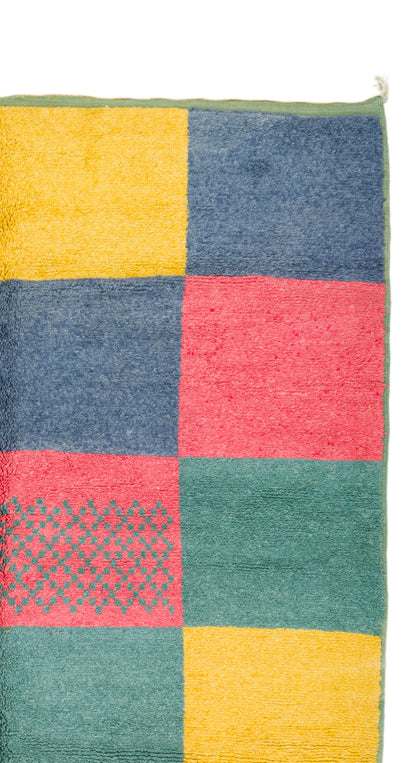A close-up of a colorful patchwork rug with large square blocks in yellow, navy blue, coral pink, and green, including one patterned green and pink block.