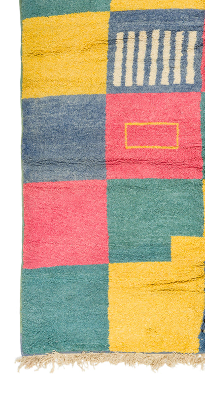 A square multicolor area rug with a patchwork design in yellow, teal, pink, and blue, featuring a fringed edge and a small geometric pattern in the top right.