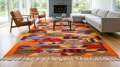 A large cubist moroccan area rug  from taznakht  with a multicolor geometric pattern and cream fringe, styled in a modern living room with a gray sectional sofa, brown leather chairs, and a glass-top coffee table.