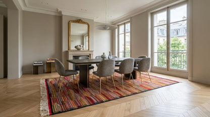 A modern dining room featuring a dark wood rectangular table, six gray upholstered chairs with gold legs, and a vibrant multicolor striped area rug from taznakht on a light wood herringbone floor. Large windows with white frames look out onto a balcony, and a gold-framed mirror hangs on the beige wall above a fireplace.